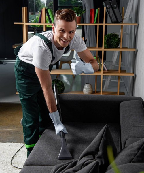 smiling young cleaning company worker cleaning sofa with vacuum cleaner and showing thumb up